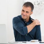 image of man with shoulder pain sitting in from of laptop on workstation