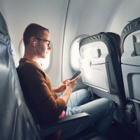 Young man (traveler) using smart phone during flight and listening music.